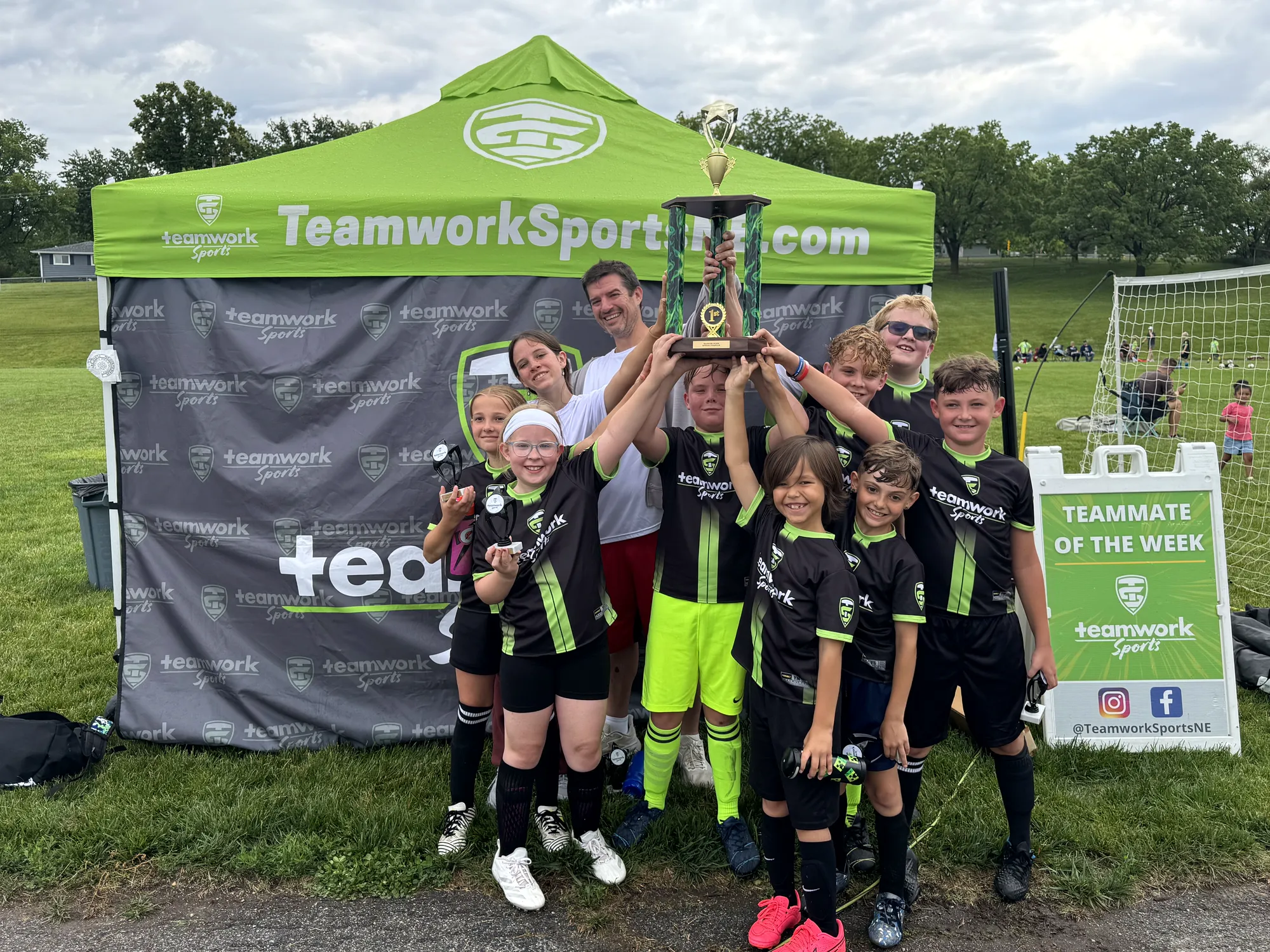A flag football team in green jerseys lifting a trophy under the Teamwork Sports tent